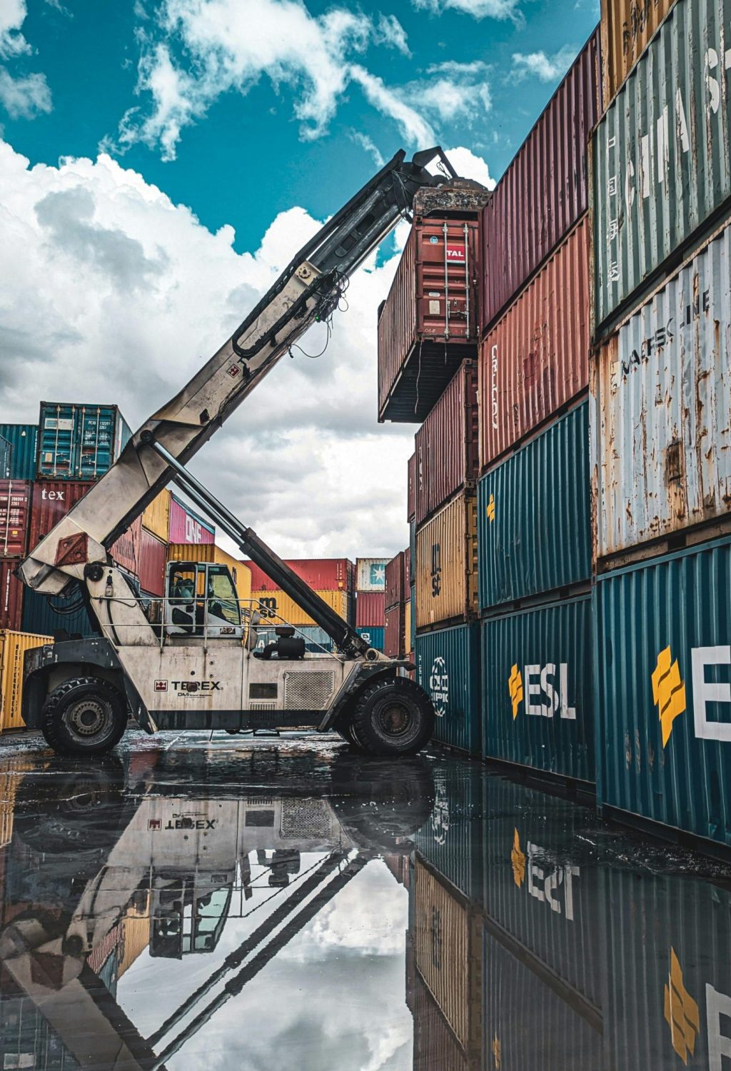 A crane loader stacking colorful shipping containers at a port with reflection in water.